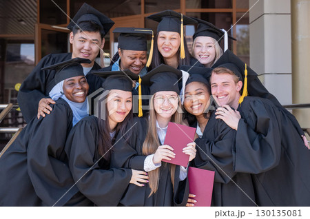 Happy students of different nationalities in graduation gowns hold their diplomas. 130135081