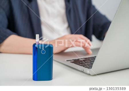 Business woman smoking an electronic cigarette while working on a laptop. Tobacco heating system in the foreground. Business woman smoking an electronic cigarette while working on a laptop. Tobacco heating system in the foreground. 130135389