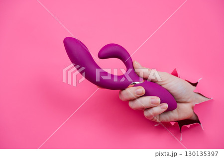 Female hand sticking out with raspberry sex toy from pink paper background. 130135397