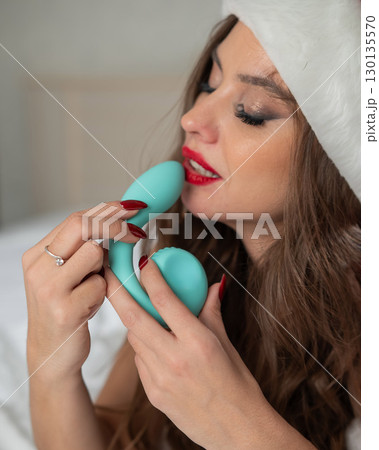 Caucasian woman in Santa Claus costume lying on bed holding curved sex toy.  130135570
