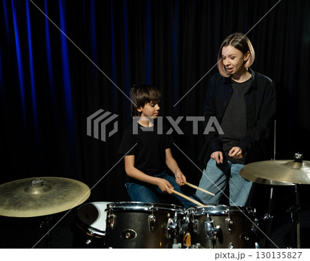 Young caucasian woman teaches a boy to play the drums in the studio on a black background. Music school student 130135827