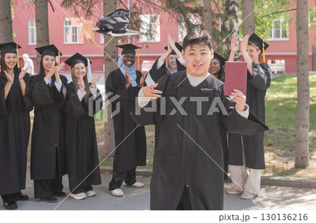 Young Asian man holds diploma while classmates applaud her. Young Asian man holds diploma while classmates applaud her. 130136216