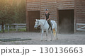 Young horsewoman riding white horse training in the paddock near wooden stable at sunset, equestrian sport, horse riding, hobby and leisure activity 130136663