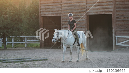 Young horsewoman riding white horse training in the paddock near wooden stable at sunset, equestrian sport, horse riding, hobby and leisure activity Young horsewoman riding white horse training in the paddock near wooden stable at sunset, equestrian sport, horse riding, hobby and leisure activity 130136663