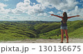 Female tourist standing with open arms, embracing the stunning landscape of rolling green hills and blue sky with clouds in Sumba Island, Indonesia, during a sunny summer day 130136675