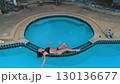 Aerial drone view of young woman in bikini relaxing on the edge of a round swimming pool with turquoise water, a cat statue is visible on the poolside 130136677