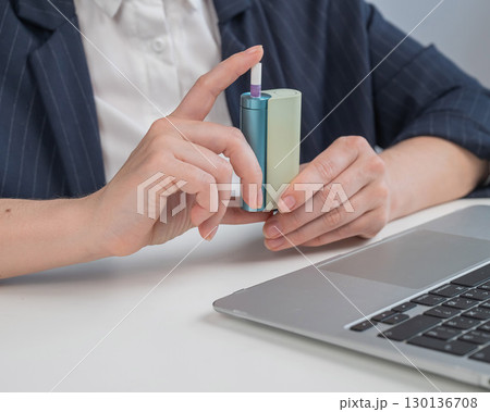 Business woman smoking an electronic cigarette while working on a laptop. Tobacco heating system.  130136708