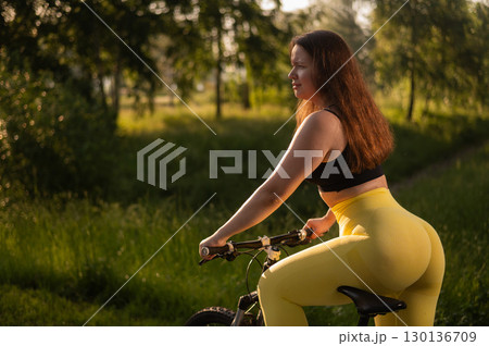 A Caucasian woman in yellow leggings and a crop top rides a bicycle in tights in the park.  130136709