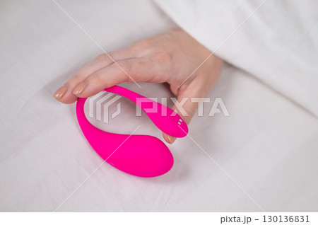 Woman holding curved pink vibrator while lying in bed. Woman holding curved pink vibrator while lying in bed. 130136831