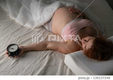 A pregnant woman lies in bed and holds an alarm clock.  130137240