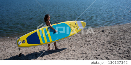 Caucasian woman walks along the beach and carries a sup board on the river in the city. Summer sport. 130137342