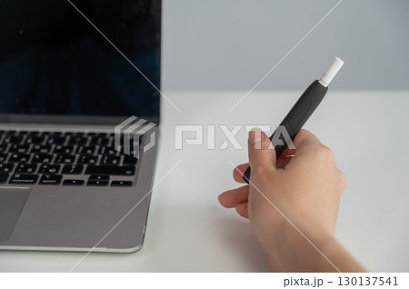 A woman smokes an electronic cigarette while working on a laptop. Tobacco heating system. A woman smokes an electronic cigarette while working on a laptop. Tobacco heating system. 130137541