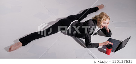 A ballerina dressed in a business suit sits in a middle twine, works at a laptop and drinks coffee. Flexible woman works at the computer 130137633