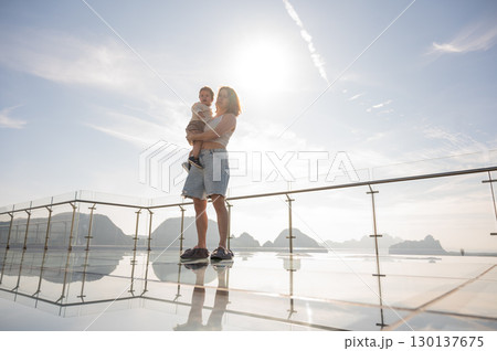 Caucasian woman holds her little son in her arms while standing on a glass bridge in the mountains.  130137675