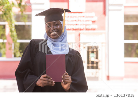 African woman in hijab and graduation gown holding diploma.  130137819