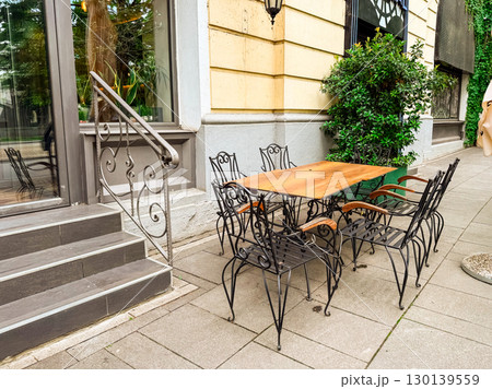 Wooden table with decorative metal chairs on cafe terrace beside window and stone stairs. Outdoor dining, hospitality, lifestyle, architecture and urban street culture. Wooden table with decorative metal chairs on cafe terrace beside window and stone stairs. Outdoor dining, hospitality, lifestyle, architecture and urban street culture. 130139559