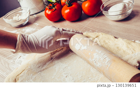 Baker kneading fresh pasta dough with rolling pin surrounded by ripe tomatoes and kitchen bowls on rustic wooden counter Baker kneading fresh pasta dough with rolling pin surrounded by ripe tomatoes and kitchen bowls on rustic wooden counter 130139651