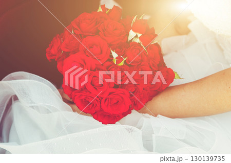Bride Holding Vibrant Red Rose Bouquet Against Flowing White Wedding Dress in Soft Golden Light 130139735
