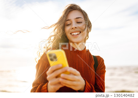 Young woman using mobile phone at beach. Cold weather. Autumn. Blogging, communication Young woman using mobile phone at beach. Cold weather. Autumn. Blogging, communication 130140482