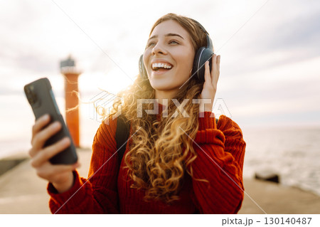Happy woman listening nice music by the waterfront while wearing headphones by a lighthouse. Happy woman listening nice music by the waterfront while wearing headphones by a lighthouse. 130140487