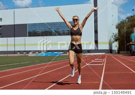 Female athlete celebrating victory on track with raised arms after hurdles race 130140491
