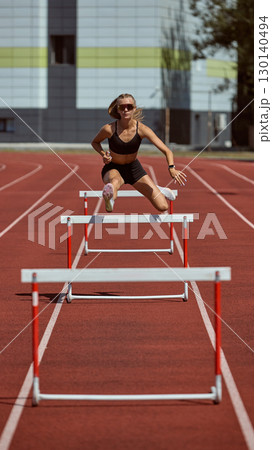 Female hurdler mid-air jump over barrier on stadium track 130140494