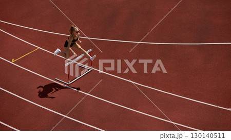 Female track athlete sprinting and clearing hurdle in powerful action motion 130140511