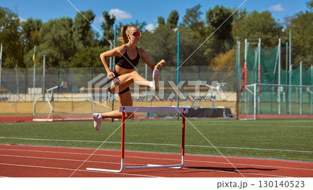Young woman hurdler jumping over barrier during outdoor training Young woman hurdler jumping over barrier during outdoor training 130140523