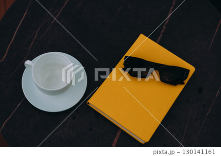 Sunglasses and a yellow book with a cup on a dark, marble table. A cup and saucer, sunglasses, and a yellow book rest on a dark, patterned surface, creating a modern, aesthetic composition. 130141161