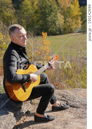 Blonde man playing string guitar outdoors in autumn forest. Concept of sound therapy, mental health and wellness rituals. Calmness tranquility audio-sensory practices. Aura farming energy 130142694