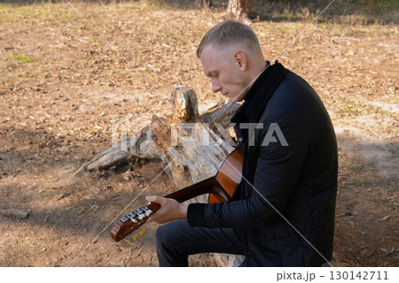 Beautiful blonde man playing acoustic guitar and singing relaxing enjoying outdoors on sunny autumn day. Artist learning and practicing playing musical instrument. Fall leaves autumnal Beautiful blonde man playing acoustic guitar and singing relaxing enjoying outdoors on sunny autumn day. Artist learning and practicing playing musical instrument. Fall leaves autumnal 130142711
