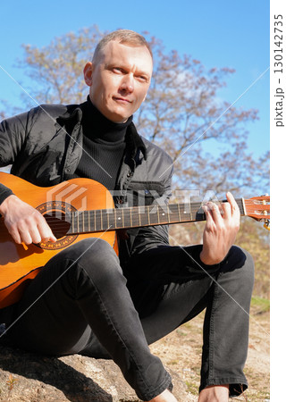 Portrait of smiling caucasian artist plays wooden acoustic guitar in autumnal park. Young songwriter plays string musical instrument hobby outside in nature fall time. Audio music healing 130142735