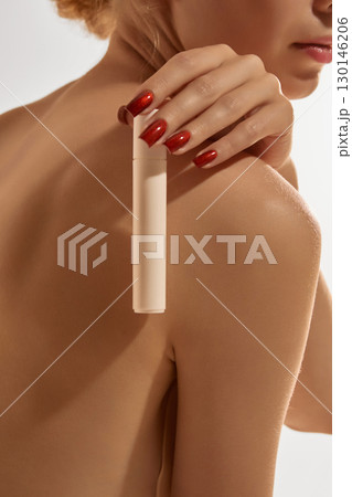 Beige cosmetic stick held by woman with red nails on bare shoulder 130146206