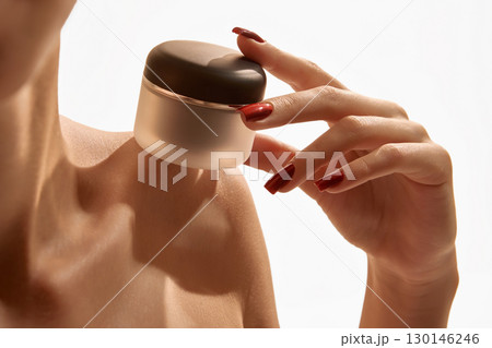 Woman hand with red nails holding frosted cream jar with dark lid on bare shoulder closeup Woman hand with red nails holding frosted cream jar with dark lid on bare shoulder closeup 130146246