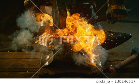 The blacksmith manually forging the molten metal on the anvil in smithy with spark fireworks 130146517