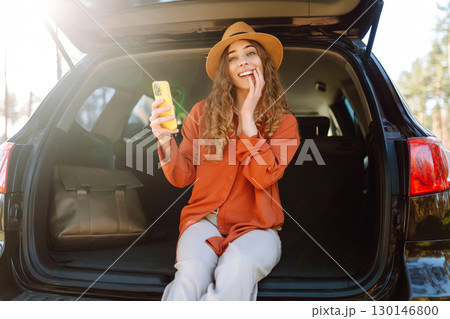 Beautiful woman sits in the trunk of a car outdoor and uses a smartphone. Lifestyle 130146800