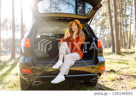 Beautiful woman sits in the trunk of a car outdoor and uses a smartphone. Lifestyle Beautiful woman sits in the trunk of a car outdoor and uses a smartphone. Lifestyle 130146803