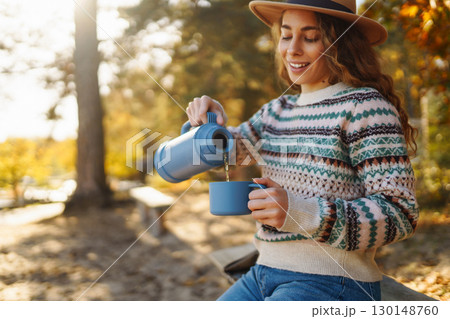 Lady drinks from thermo mug of coffee in nature. Warm up in sunny autumn forest. Travel, adventure. 130148760