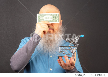 bald man with a large white beard holds a dollar and a model of a grocery basket 130149004