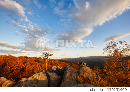 Golden hour bathes autumn forest in warm light, highlighting vibrant red and orange foliage. Majestic rock formations, under sky with scattered clouds. Dovbush Rocks, Carpathian mountains, Ukraine. Golden hour bathes autumn forest in warm light, highlighting vibrant red and orange foliage. Majestic rock formations, under sky with scattered clouds. Dovbush Rocks, Carpathian mountains, Ukraine. 130151469
