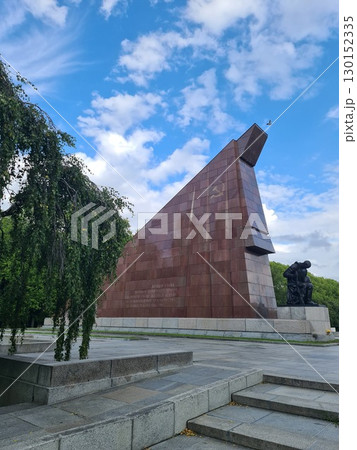 Berlin, Germany - August 1, 2025: Portal of the Soviet War Memorial in Treptower Park consisting of a pair of stylized Soviet flags built of red granite flanked by two statues of kneeling soldiers Berlin, Germany - August 1, 2025: Portal of the Soviet War Memorial in Treptower Park consisting of a pair of stylized Soviet flags built of red granite flanked by two statues of kneeling soldiers 130152335
