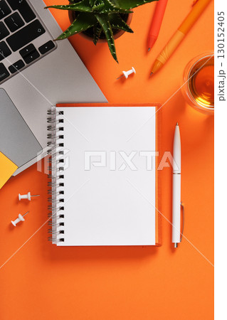 Blank notebook with laptop keyboard, pens, and cup of tea on orange desk. Workspace for study, planning, and creativity. 130152405