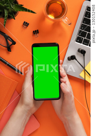 Hands holding smartphone with green screen. Laptop, tea, and stationery on orange desk. Place for your app design or screenshot. 130152408