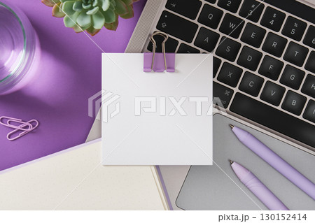 Blank note with binder clip on laptop keyboard with pens, notebook, and plant on purple desk. Copyspace. 130152414
