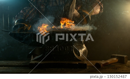Close-up of blacksmith forging glowing hot iron on anvil with flying sparks 130152487