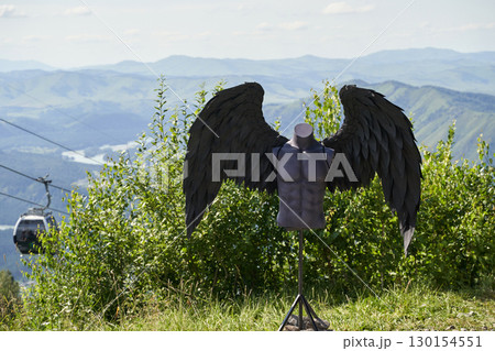 Black wings on mannequin against mountain landscape with blue sky and cable car 130154551