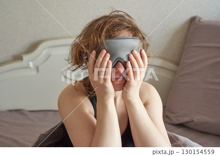 Caucasian female adult waking up in bed wearing sleep mask, hands on face 130154559