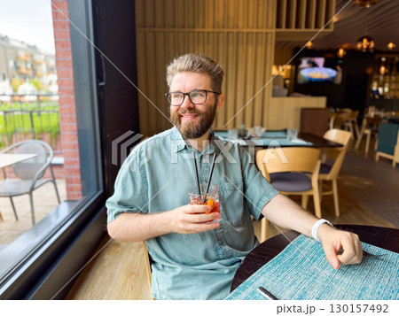Man drinking cocktail with straw while sitting near restaurant window. Refreshment, leisure, and hospitality lifestyle captured in casual dining environment. Man drinking cocktail with straw while sitting near restaurant window. Refreshment, leisure, and hospitality lifestyle captured in casual dining environment. 130157492