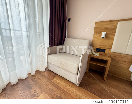 Light beige armchair near window and balcony in a modern hotel room. Comfort, rest, and hospitality interior design for leisure, travel, and accommodation experience. 130157515