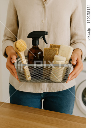 Person holding stylish mesh basket filled with eco friendly cleaning supplies wooden brush glass spray bottle and natural sponges, conveying sense of care and cleanliness 130158184